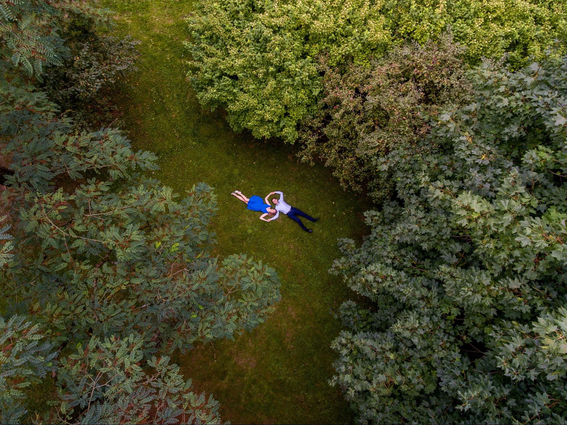 Para leżąca na trawie w parku w Słupsku, widziana z lotu ptaka. Kobieta w niebieskiej sukience, mężczyzna w białej koszuli. Otoczenie: drzewa i trawa.