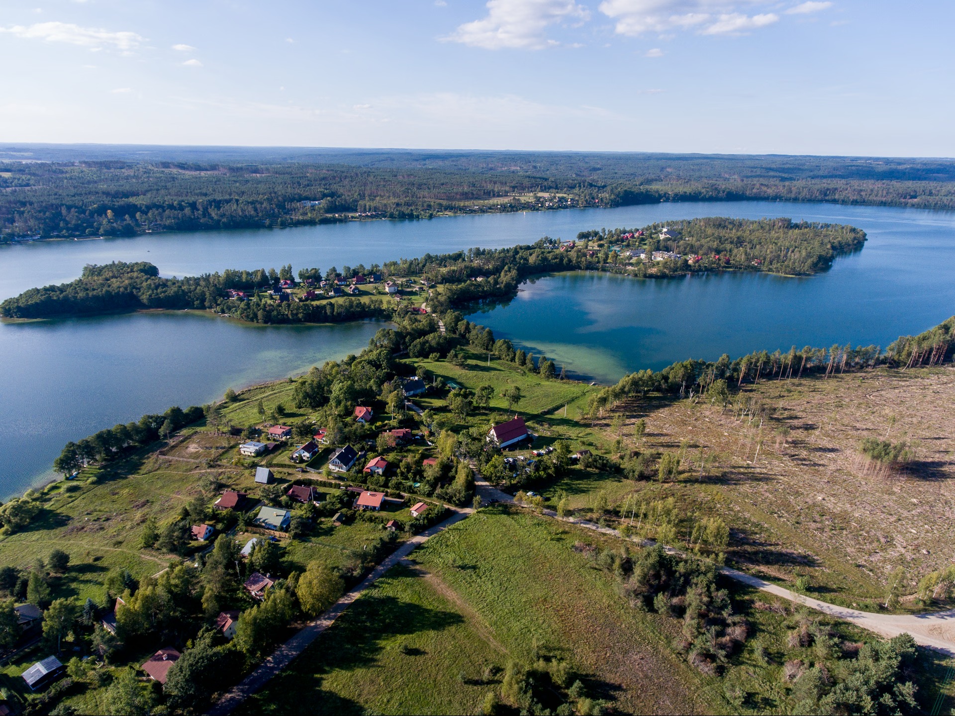 Panoramiczny widok z lotu ptaka na malownicze jezioro z wyspami i zabudowaniami, otoczone lasami i polami w słoneczny dzień. Ujęcie z góry ukazuje detale krajobrazu.