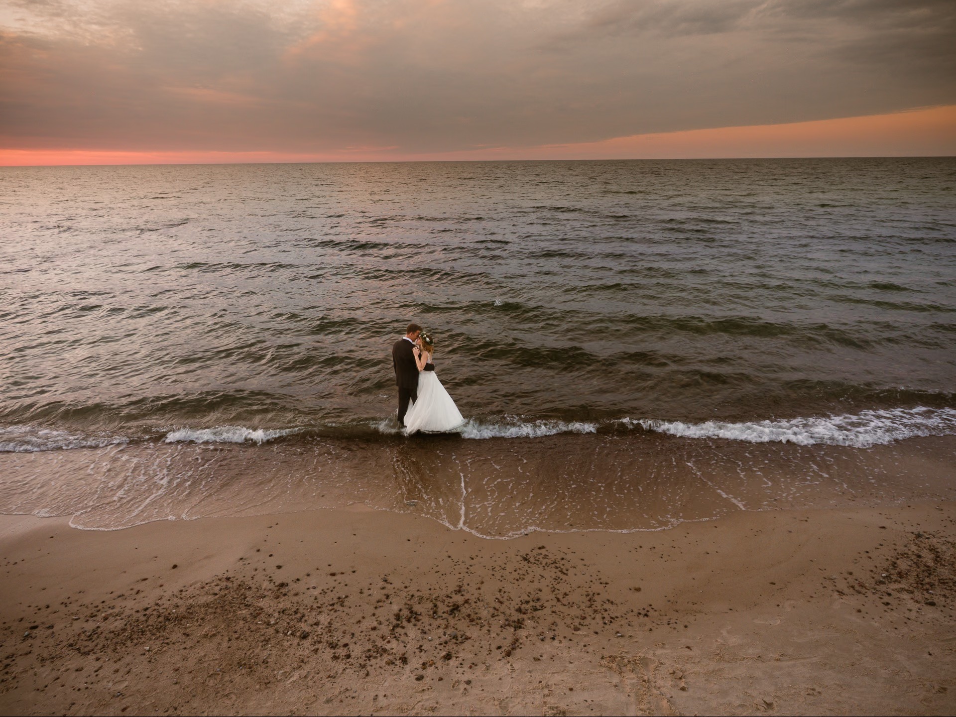 Para młoda w objęciach stoi w wodzie na plaży o zachodzie słońca. Delikatne fale obmywają brzeg, a niebo przybiera ciepłe, pomarańczowe i różowe barwy.