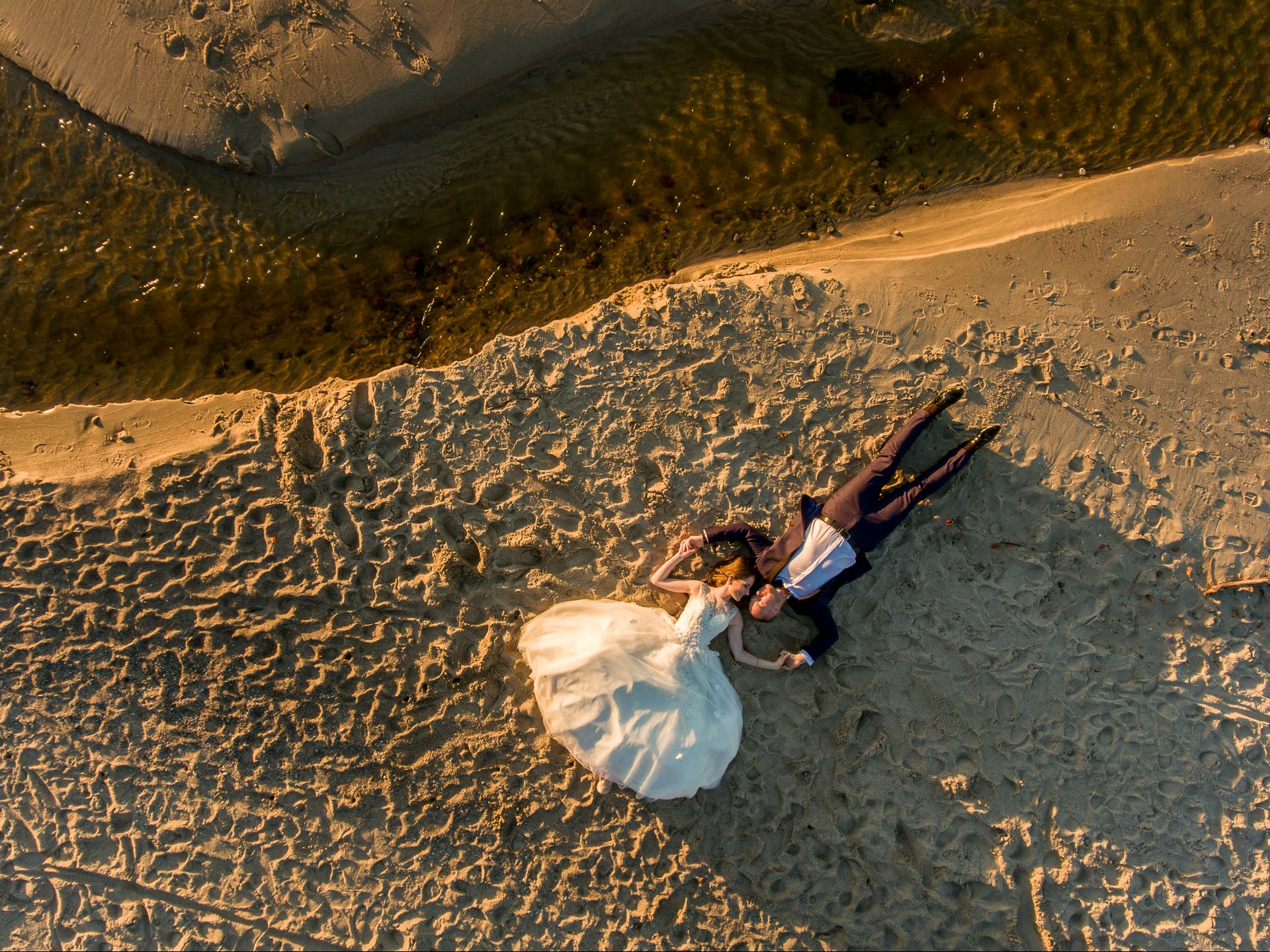 Zdjęcie z drona: para młoda leży na piasku obok rzeki, ona w białej sukni, on w granatowym garniturze. Ujęcie z góry, ciepłe światło zachodzącego słońca.