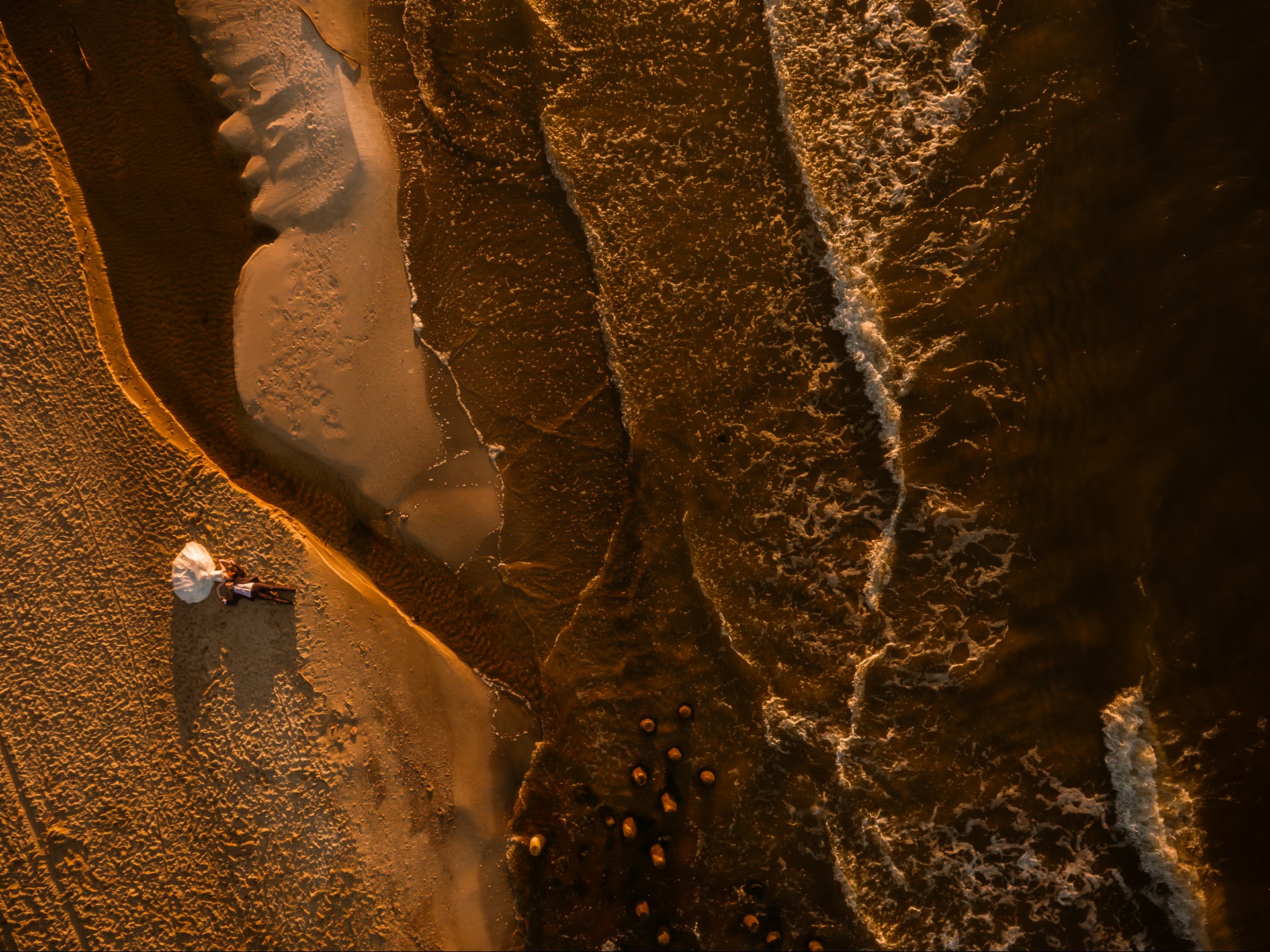 Zdjęcie z drona: para młoda leży na piaszczystej plaży, obok woda z falami. Ciepłe, złote światło zachodzącego słońca. Ujęcie z góry, podkreślające przestrzeń i intymność.