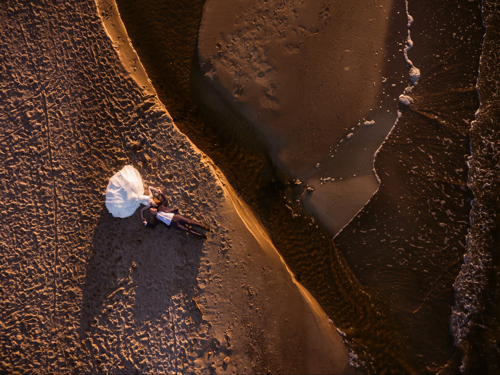 Para młoda leży na piasku obok rzeki, widok z drona. Panna młoda w białej sukni, pan młody w garniturze. Ciepłe światło zachodzącego słońca.