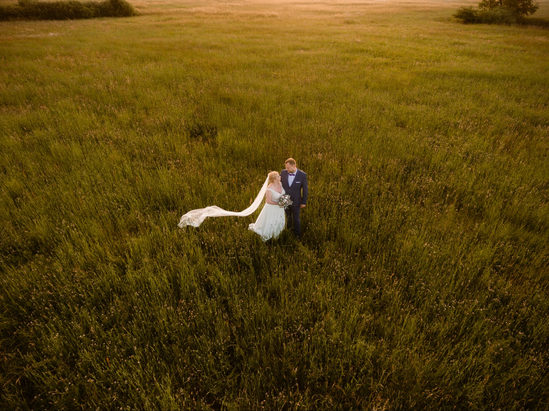 Para młoda w plenerze, ujęcie z drona. Panna młoda w białej sukni i welonie, pan młody w granatowym garniturze. W tle rozległe pole trawy w ciepłych, zachodzących promieniach słońca.