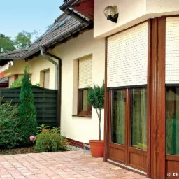 Zamknięte kremowe osłony okienne zewnętrzne na parterze domu z drewnianą stolarką okienną, widoczne z zewnątrz na tle kostki brukowej i ogrodowej zieleni.