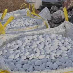 Białe otoczaki w workach typu Big Bag z żółtymi uchwytami, przygotowane do transportu. W tle kamienie i żwir.