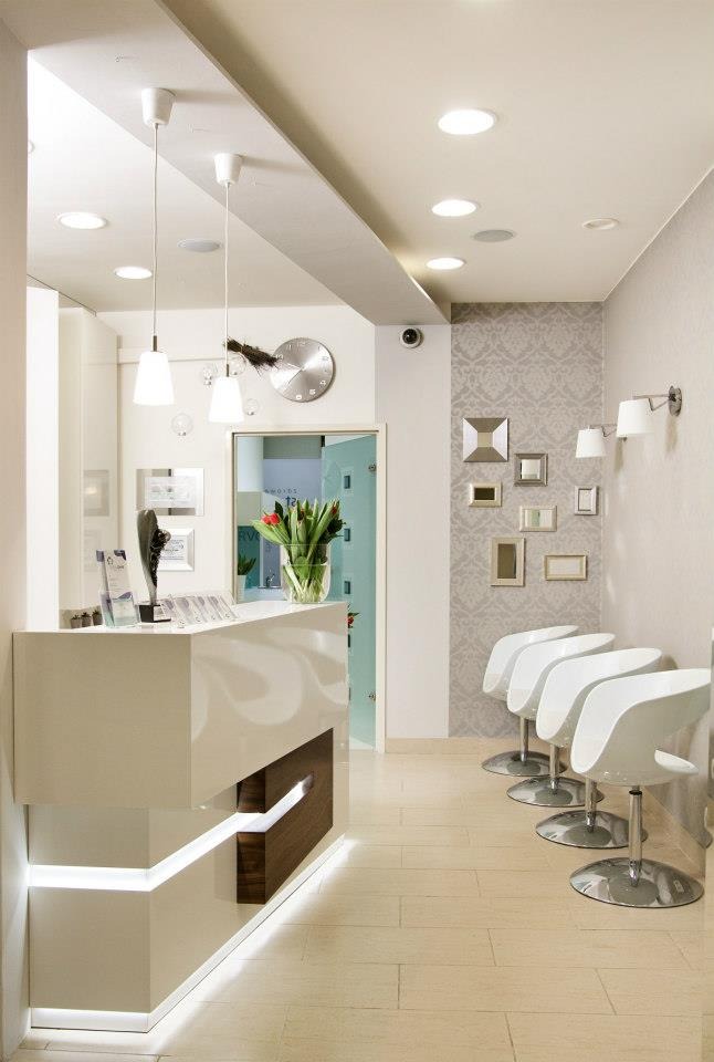 Nowoczesne wnętrze biura z białą recepcją z podświetleniem LED, czterema białymi krzesłami obrotowymi i ścianą z dekoracyjną tapetą oraz ramkami.