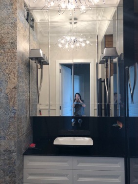 Nowoczesna łazienka z dużym lustrem podzielonym na panele, podwieszana umywalka nablatowa na czarnym blacie, białe szafki pod umywalką, kinkiety po bokach lustra, dekoracyjna lampa sufitowa...