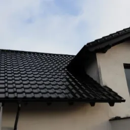 Fragment domu z ciemną dachówką karpiówką i jasną elewacją, widziany z dołu pod kątem. Widoczne okno na poddaszu i fragment nieba.