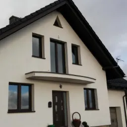 Dom jednorodzinny z białą elewacją i ciemną stolarką okienną. Widoczny fragment dachu, balkon nad wejściem i antena na dachu sąsiedniego budynku.