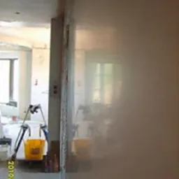 Świeżo położona, gładka, jasna ściana w trakcie schnięcia, widoczne refleksy światła, w tle drabina malarska i żółte wiaderko.