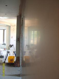 Świeżo położona, gładka, jasna ściana w trakcie schnięcia, widoczne refleksy światła, w tle drabina malarska i żółte wiaderko.