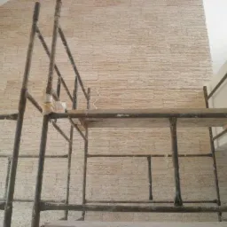Ściana z dekoracyjną, beżową okładziną kamienną wewnątrz budynku, widoczna z poziomu rusztowania. Częściowe ujęcie sufitu i fragmentu ściany pomalowanej na biało.