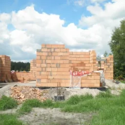Budowa domu w trakcie: wzniesione ściany z czerwonej cegły, widoczne zbrojenie fundamentów, teren budowy z zielenią w tle, błękitne niebo z chmurami.