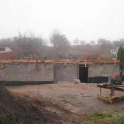 Budowa domu w trakcie: ściany z bloczków, szalunki drewniane, teren budowy w mglisty dzień. Widoczny stół roboczy z narzędziami.