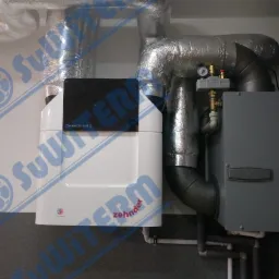 Biała jednostka centralna z czarnym panelem Zehnder ComfoAir Q, podłączona do izolowanych przewodów wentylacyjnych, zamontowana na ścianie z szarą dolną częścią, widoczny manometr.