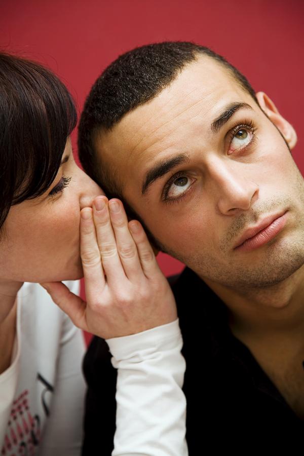 Kobieta szepcze coś do ucha zamyślonemu mężczyźnie na czerwonym tle, być może przekazując sekret lub plotkę.