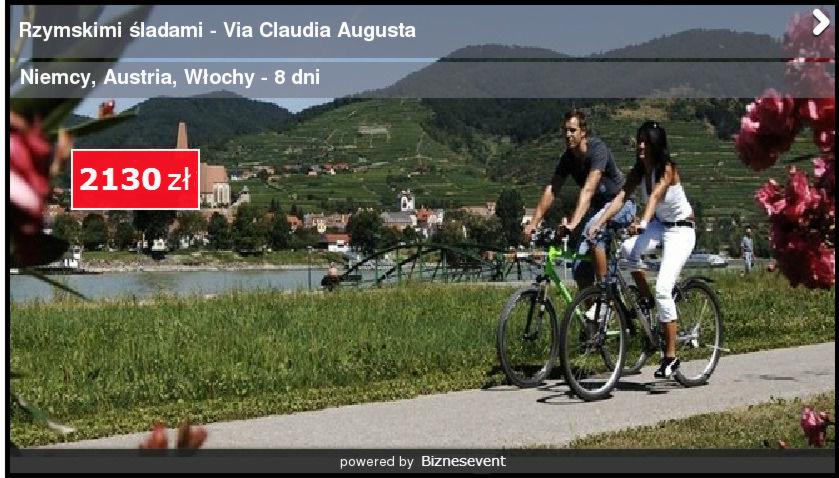 Para rowerzystów na ścieżce rowerowej wzdłuż rzeki, w tle malownicza miejscowość z kościołem i winnicami na wzgórzach, kwiaty w prawym rogu kadru.