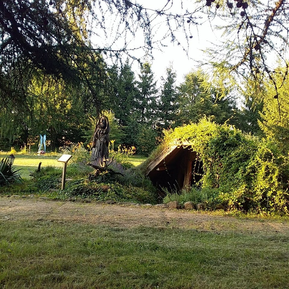 Ogród z drewnianą rzeźbą kobiety i wejściem do ziemianki porośniętej zielenią, w tle drzewa iglaste i postać w niebieskim stroju.
