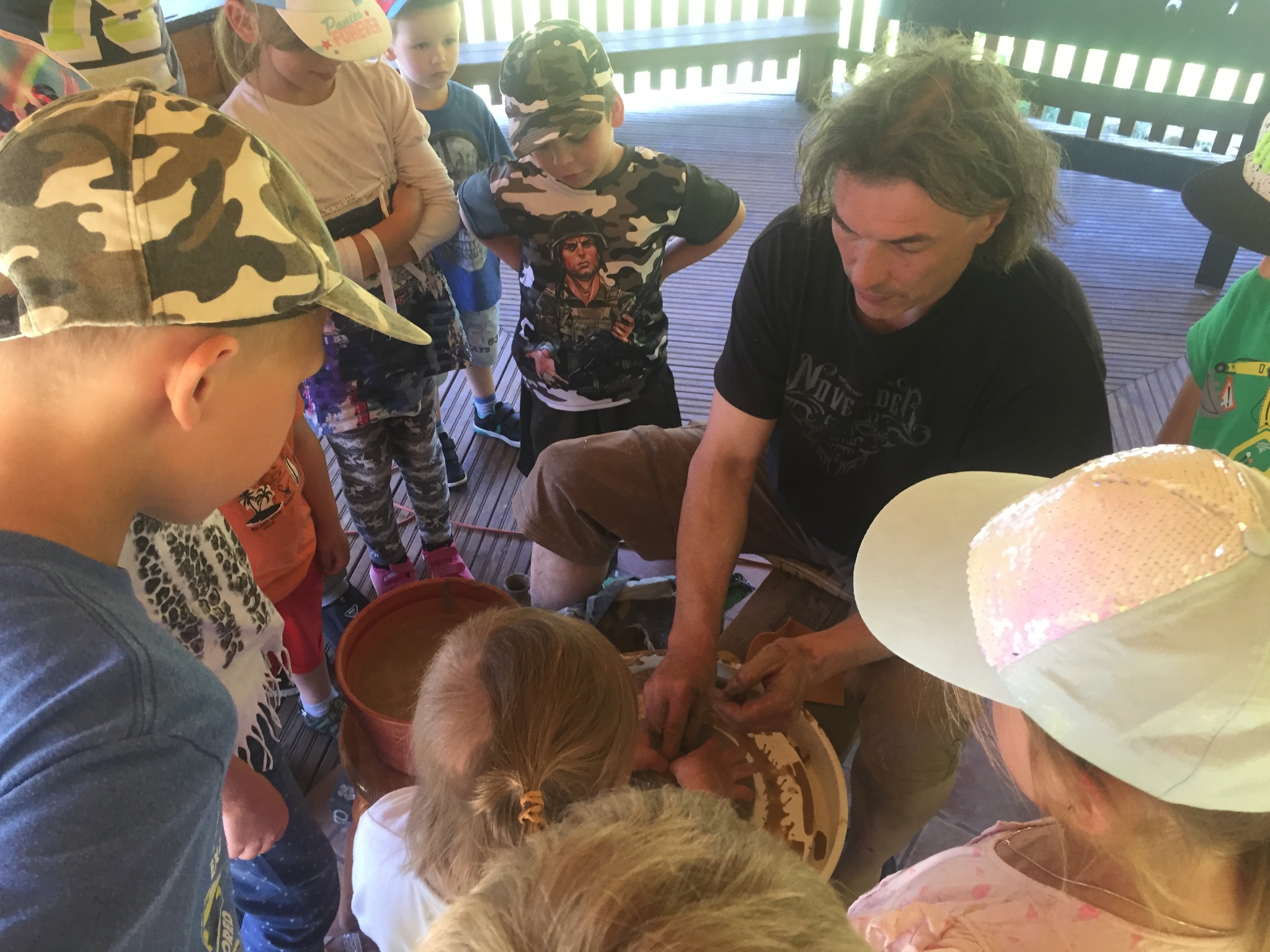 Warsztaty garncarskie: mężczyzna formuje glinę na kole garncarskim w otoczeniu grupy dzieci, obserwujących proces.
