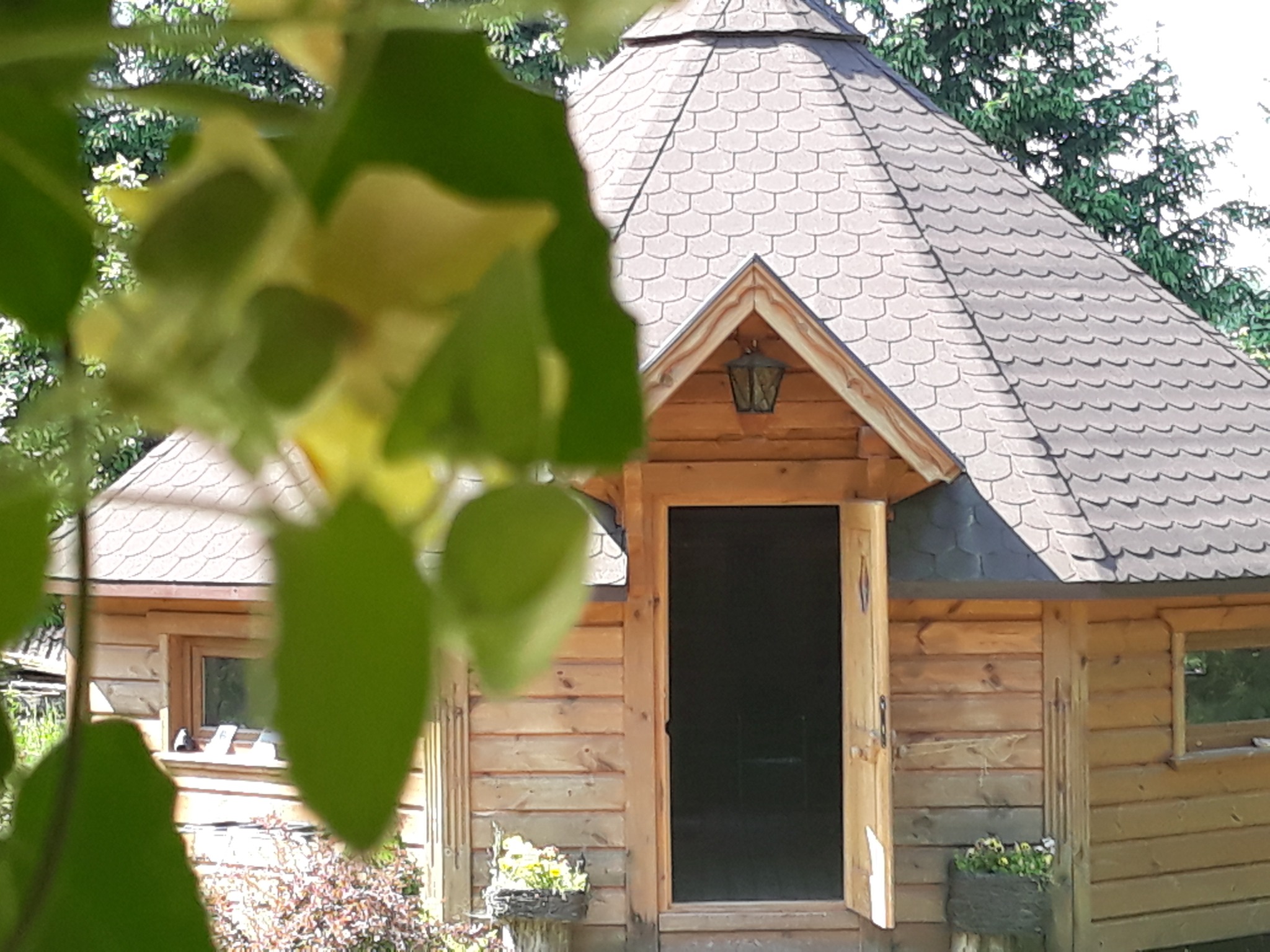 Drewniana altana ogrodowa z otwartymi drzwiami i spadzistym dachem pokrytym gontem bitumicznym, widoczna przez liście drzewa, z donicami kwiatów po bokach wejścia.