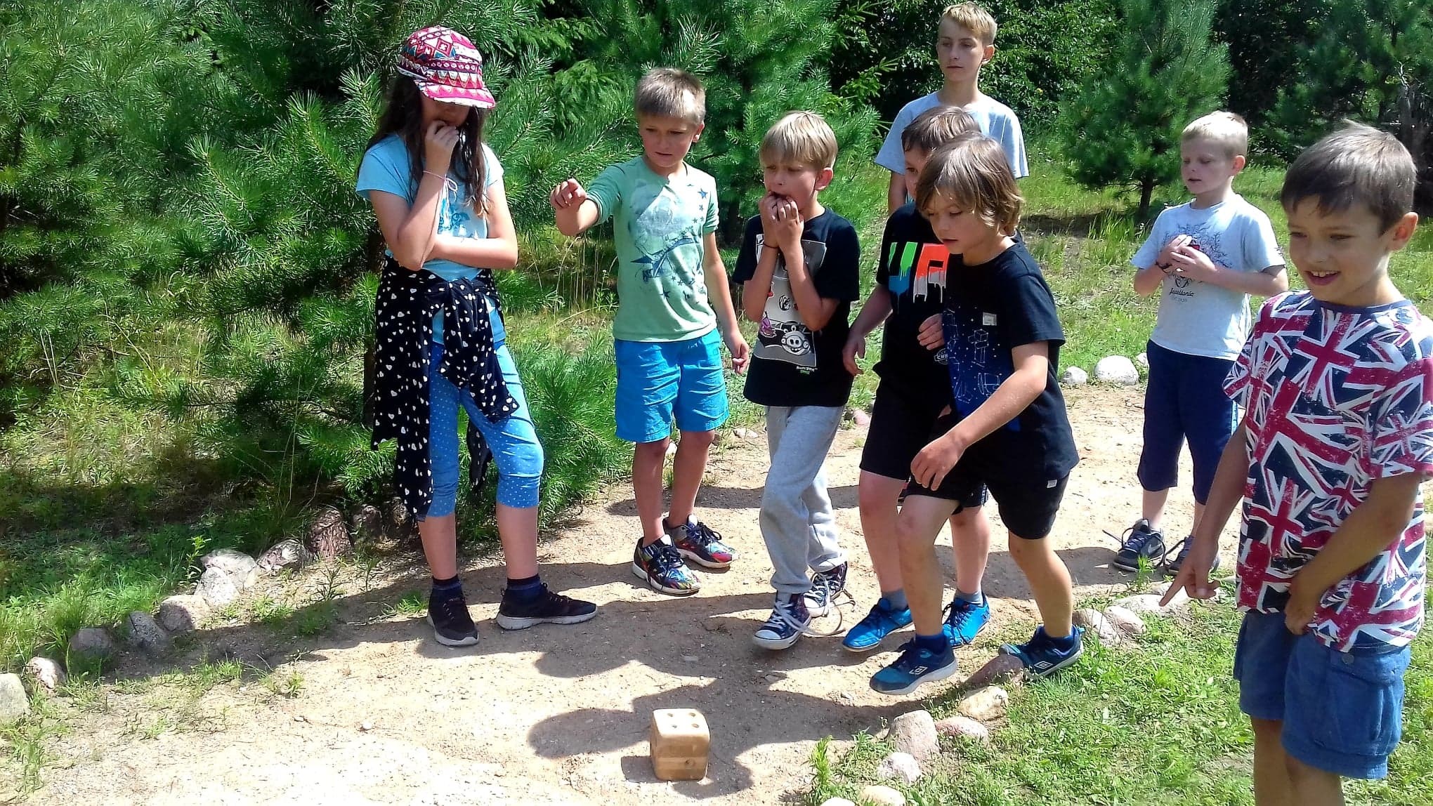 Grupa dzieci w różnym wieku, stojąca wokół drewnianej kostki na ziemi podczas gry terenowej na świeżym powietrzu, otoczona drzewami i zielenią.