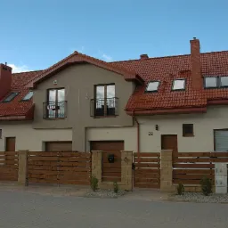 Nowoczesne, poziome ogrodzenie drewniane z elementami z piaskowca, otaczające szereg domów z czerwoną dachówką i balkonami z czarnymi balustradami.