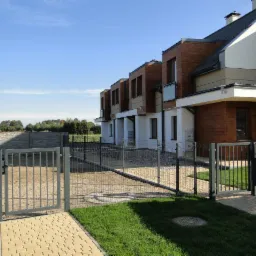 Szara metalowa furtka zamykająca wejście na posesję szeregowca, w tle betonowe ogrodzenie i zieleń.