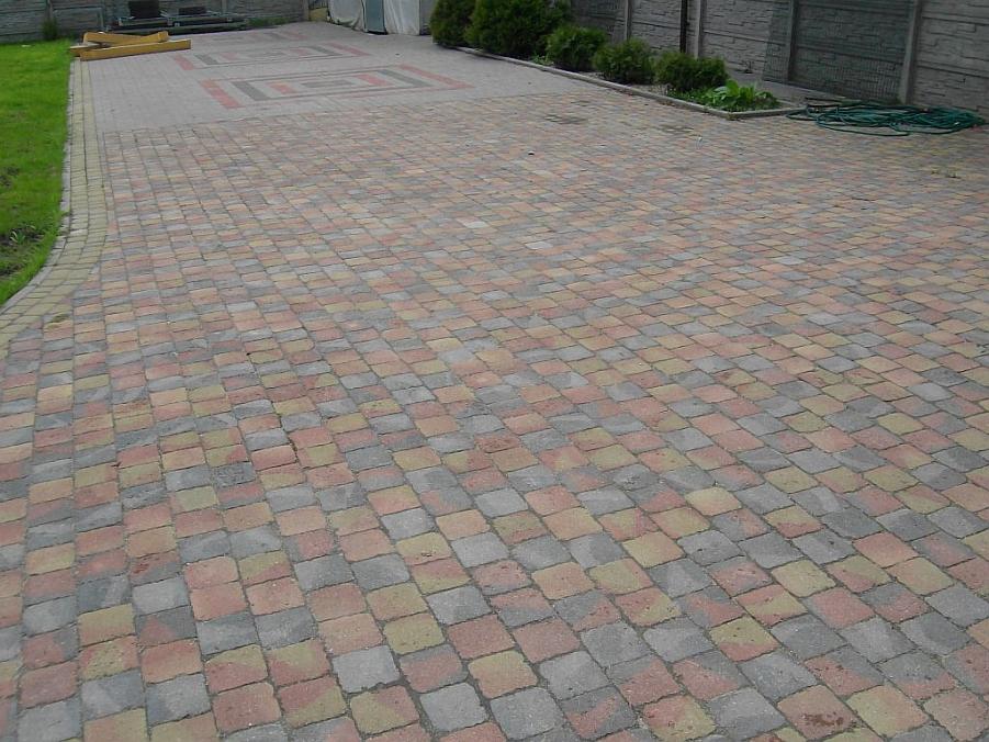 Szeroki podjazd wyłożony wielokolorową kostką brukową w odcieniach czerwieni, szarości i żółci, z ozdobnym wzorem w centralnej części, otoczony zielonym trawnikiem i betonowym ogrodzeniem.