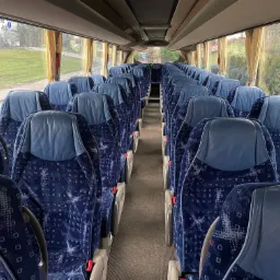 Wnętrze autobusu turystycznego z rzędami niebieskich foteli z wzorzystą tapicerką i widokiem na krajobraz za oknem.