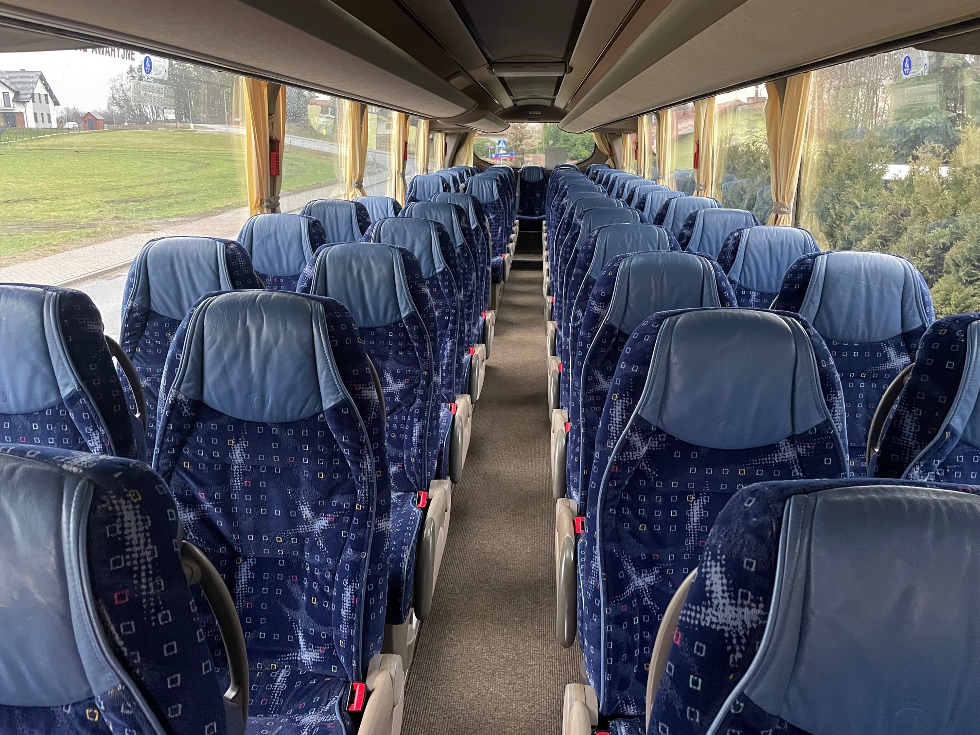 Wnętrze autobusu turystycznego z rzędami niebieskich foteli z wzorzystą tapicerką i widokiem na krajobraz za oknem.