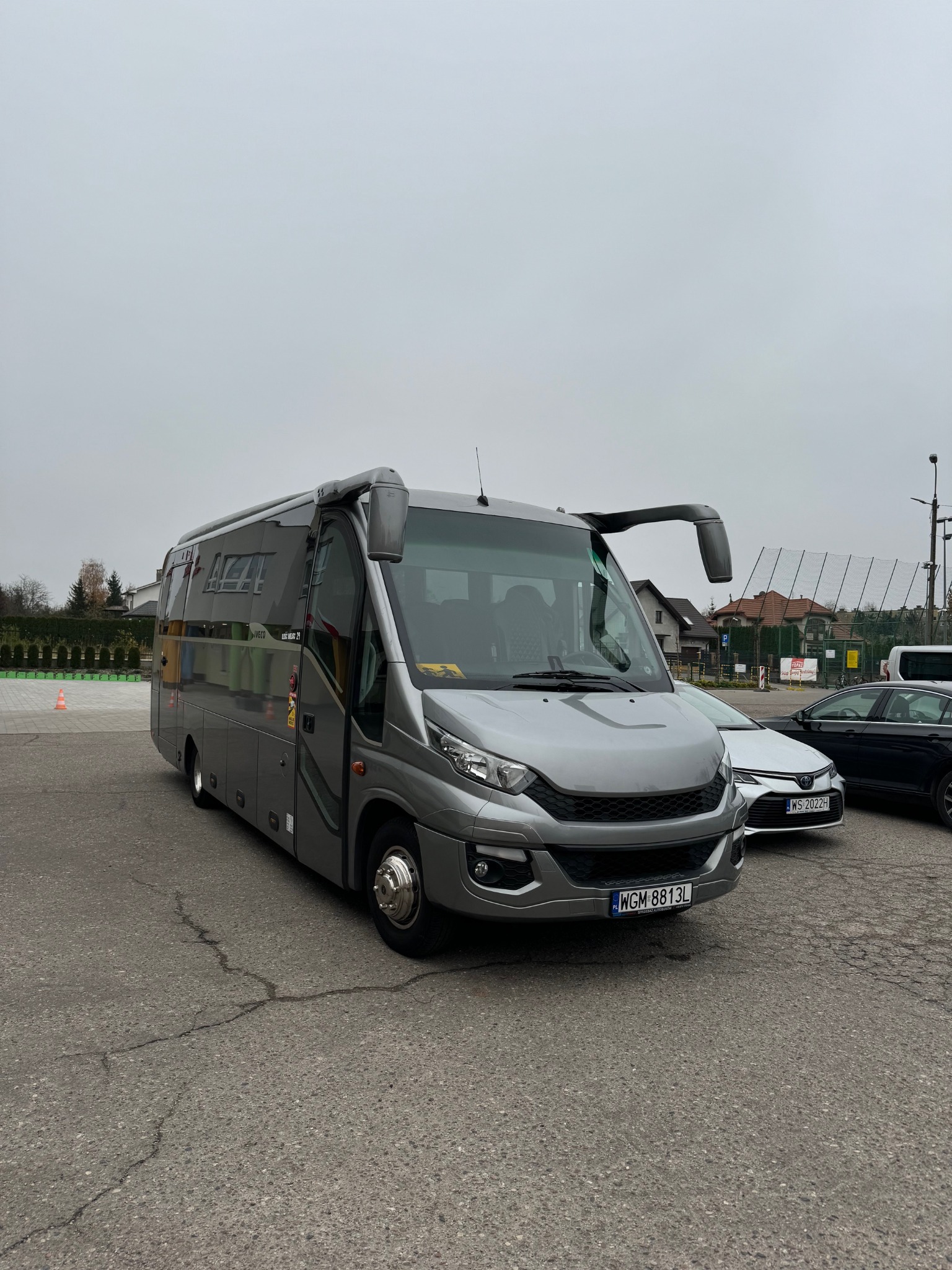 Szary minibus Iveco z numerem rejestracyjnym WGM 8813L zaparkowany na parkingu obok innych samochodów w pochmurny dzień.