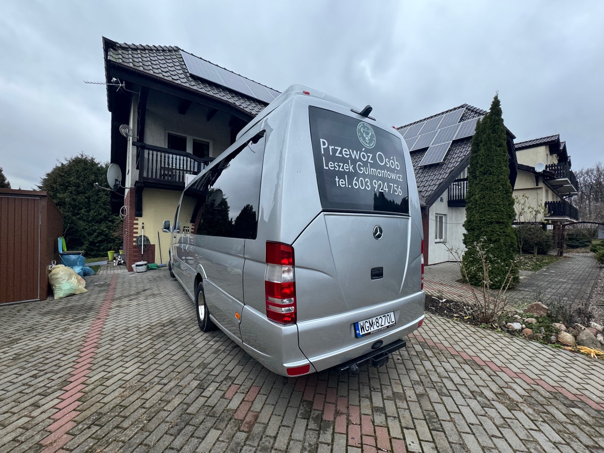Srebrny bus Mercedes z napisem 'Przewóz Osób Leszek Gulmantowicz tel. 603 924 756' na tylnej szybie, zaparkowany na brukowanym podjeździe przed domem z panelami słonecznymi na dachu.