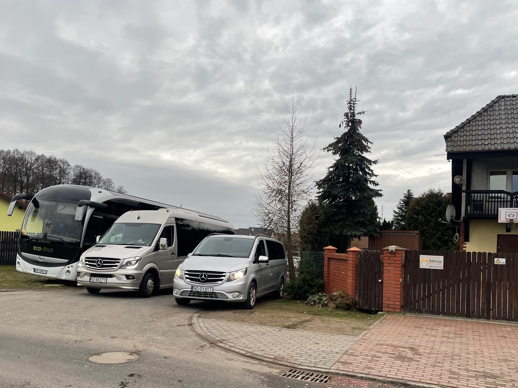 Flota pojazdów: autokar Eco-bus i dwa srebrne busy Mercedes, zaparkowane przed posesją z ceglanym ogrodzeniem i domem z balkonem.