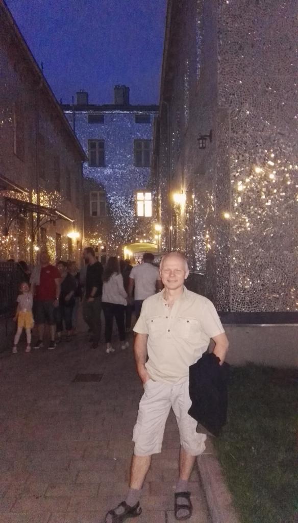 Mężczyzna w jasnej koszuli i spodenkach stoi na tle uliczki z budynkami pokrytymi mozaiką, w tle przechodzą ludzie, wieczorne oświetlenie.