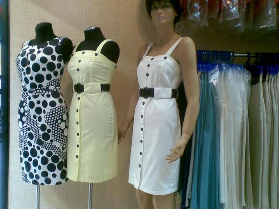 Wystawa trzech letnich sukienek: czarno-biała w grochy, jasnożółta i biała, każda z czarnym paskiem, prezentowane na manekinach w szwalni, z widocznymi wiszącymi ubraniami w tle.