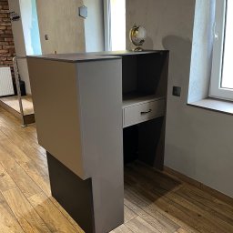 ArMebel - Nowoczesna, szara szafka z wysuwanym blatem, globusem na górze i szufladą, na tle drewnianej podłogi i okna. Minimalistyczny design.