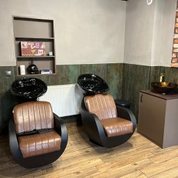 ArMebel - Dwa fotele fryzjerskie z umywalkami w salonie, brązowe obicia, czarne konstrukcje, drewniana podłoga, ściana z cegły i półka z dekoracjami w tle.