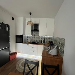 ArMebel - Biała kuchnia na wymiar z drewnianym blatem, czarnym piekarnikiem i lodówką. Nad blatem wzorzyste płytki, a pod szafkami ledowe oświetlenie. W tle widoczny okrągły dywan.