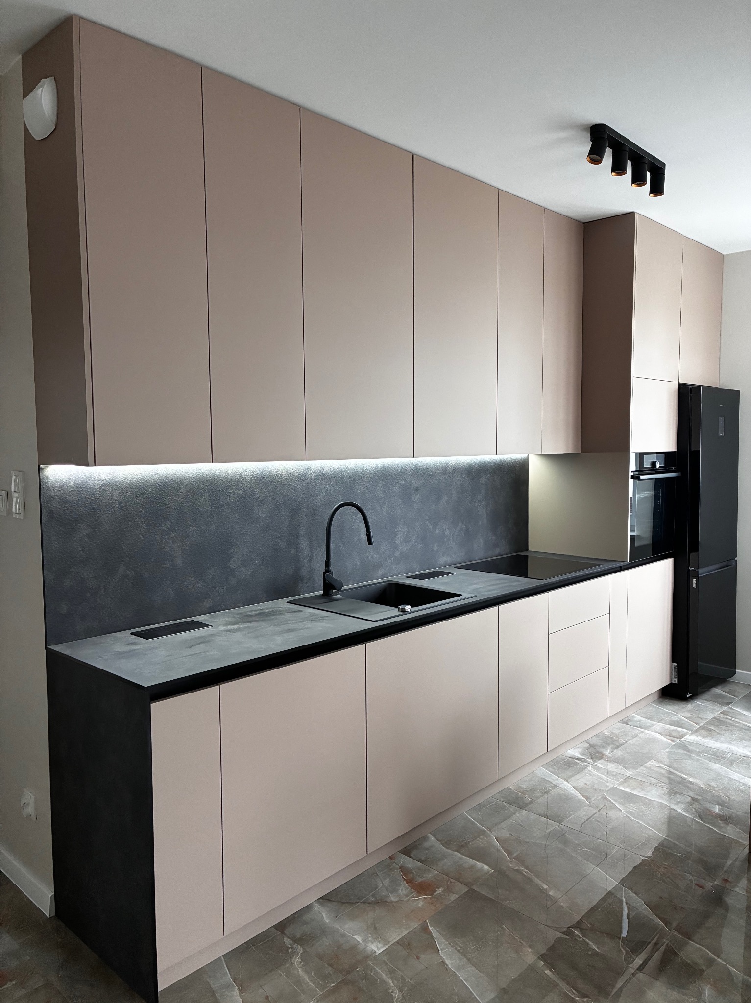 Nowoczesna kuchnia z beżowymi frontami szafek, czarnym blatem i sprzętem AGD. Podświetlenie LED nad blatem roboczym. Marmurowe płytki podłogowe.