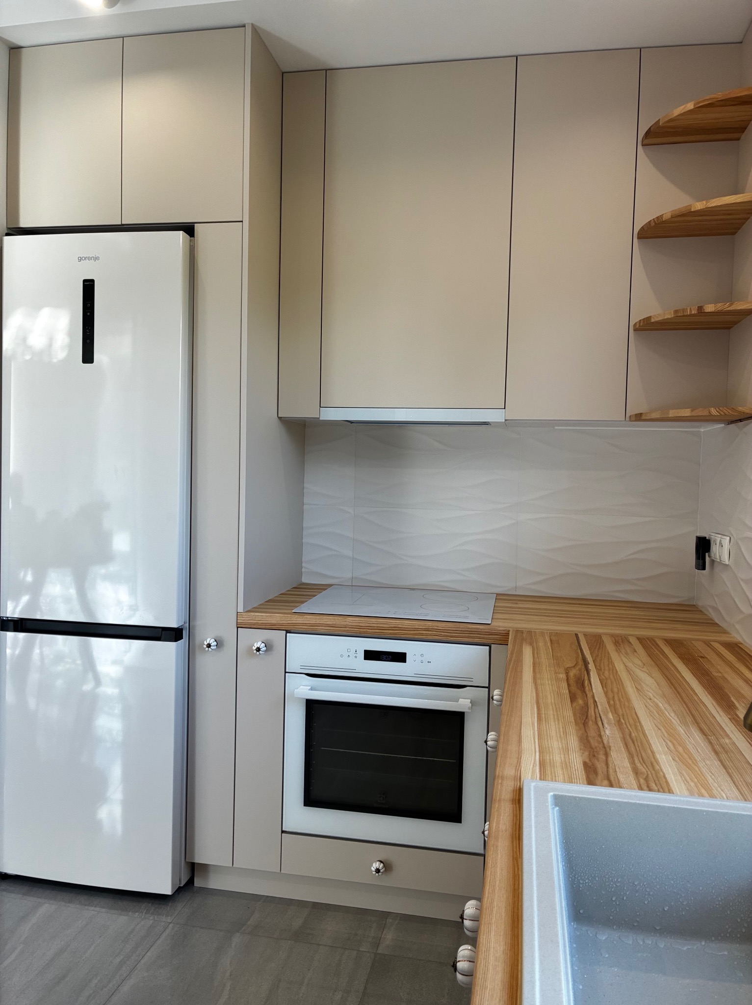 Jasna kuchnia na wymiar z drewnianym blatem i półkami w Miłomłynie. Widoczna zabudowa z lodówką i piekarnikiem, minimalistyczny design.