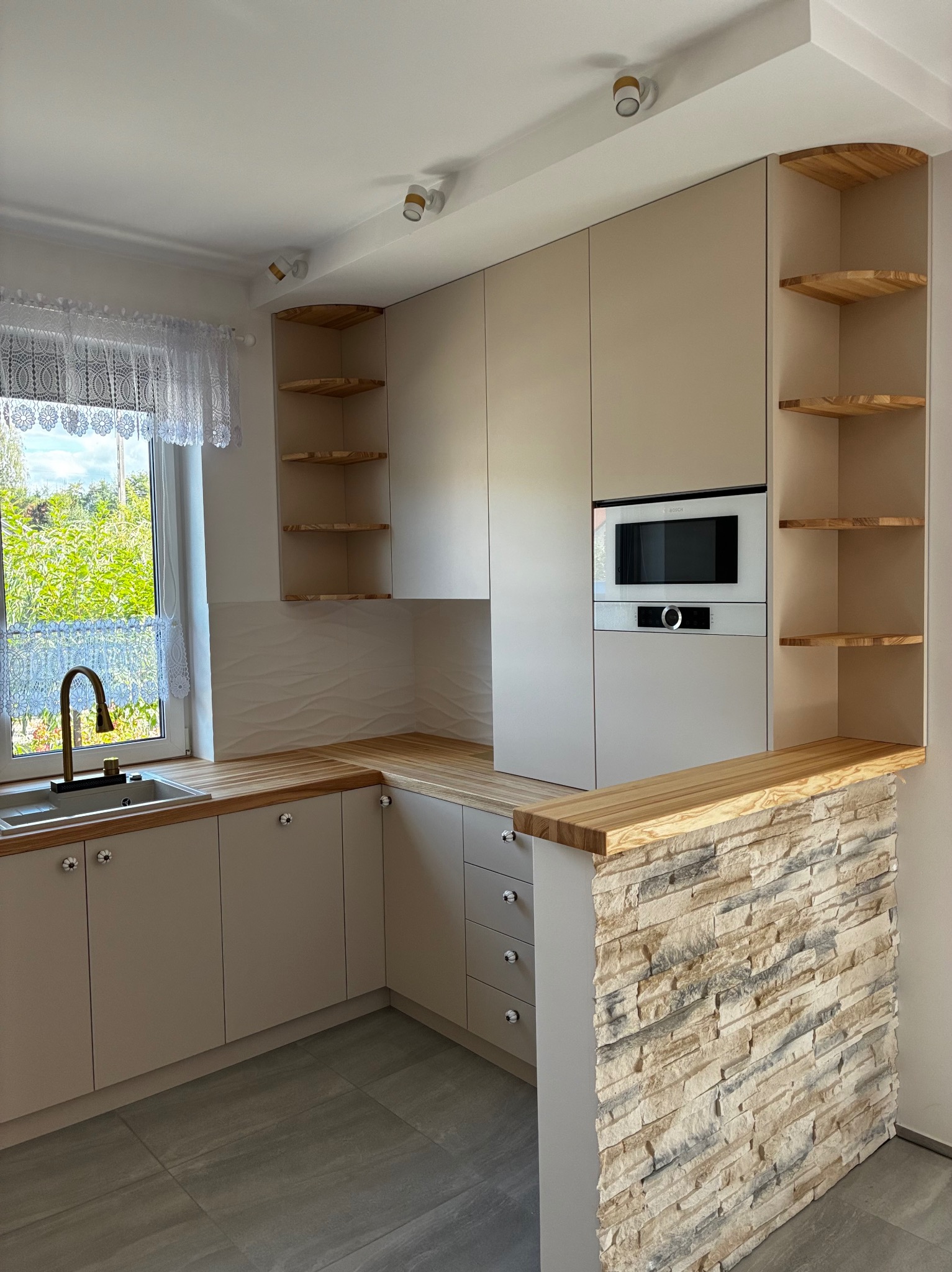 Jasna kuchnia z drewnianym blatem i barkiem wykończonym kamieniem dekoracyjnym. Widoczne otwarte półki i nowoczesny piekarnik w zabudowie. Zlewozmywak pod oknem z koronkową firanką.