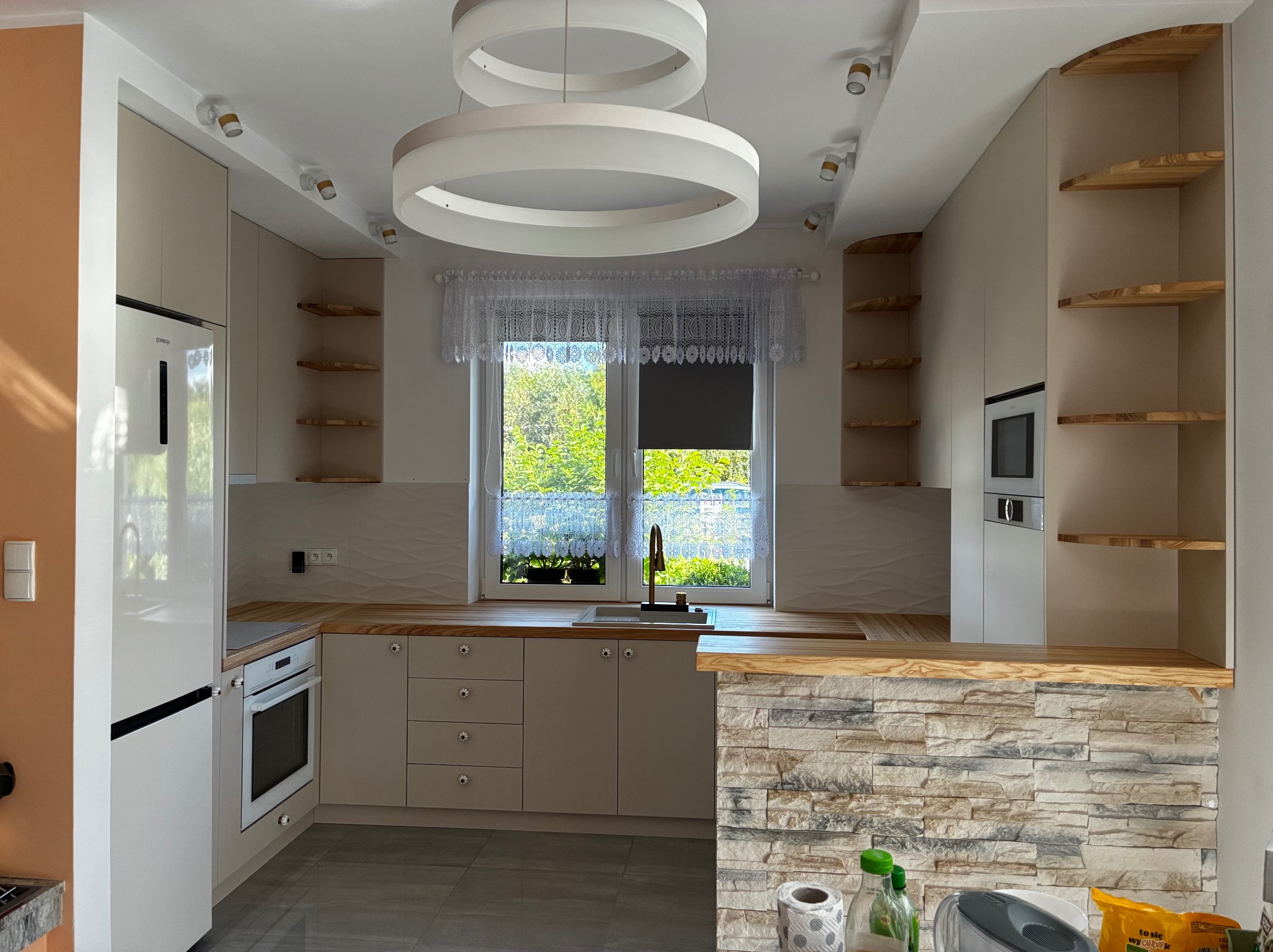 Jasna kuchnia z drewnianymi blatami i otwartymi półkami, oświetlona nowoczesną lampą pierścieniową. Ściana z dekoracyjnej cegły dodaje charakteru.