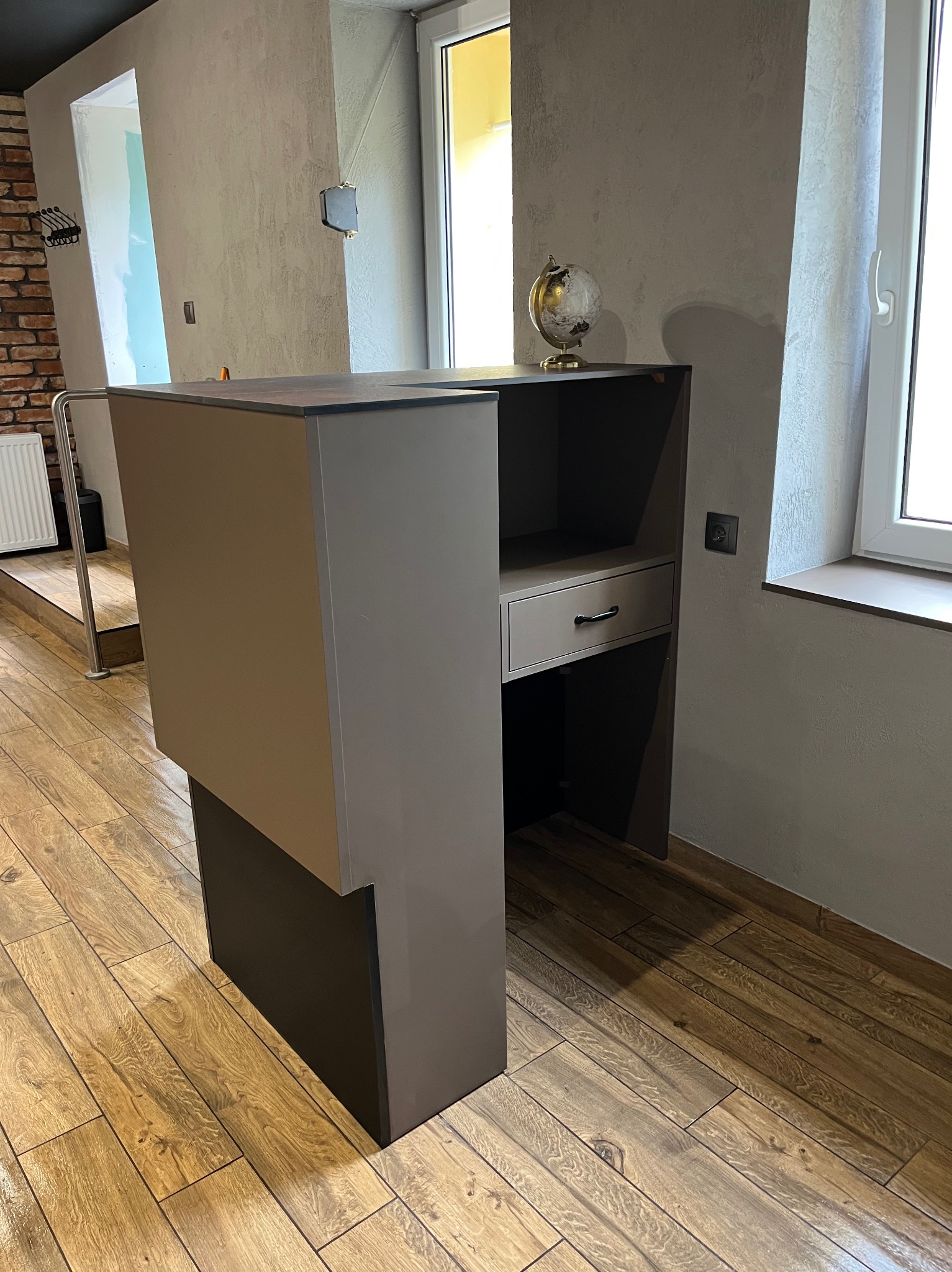 Nowoczesna, szara szafka z wysuwanym blatem, globusem na górze i szufladą, na tle drewnianej podłogi i okna. Minimalistyczny design.