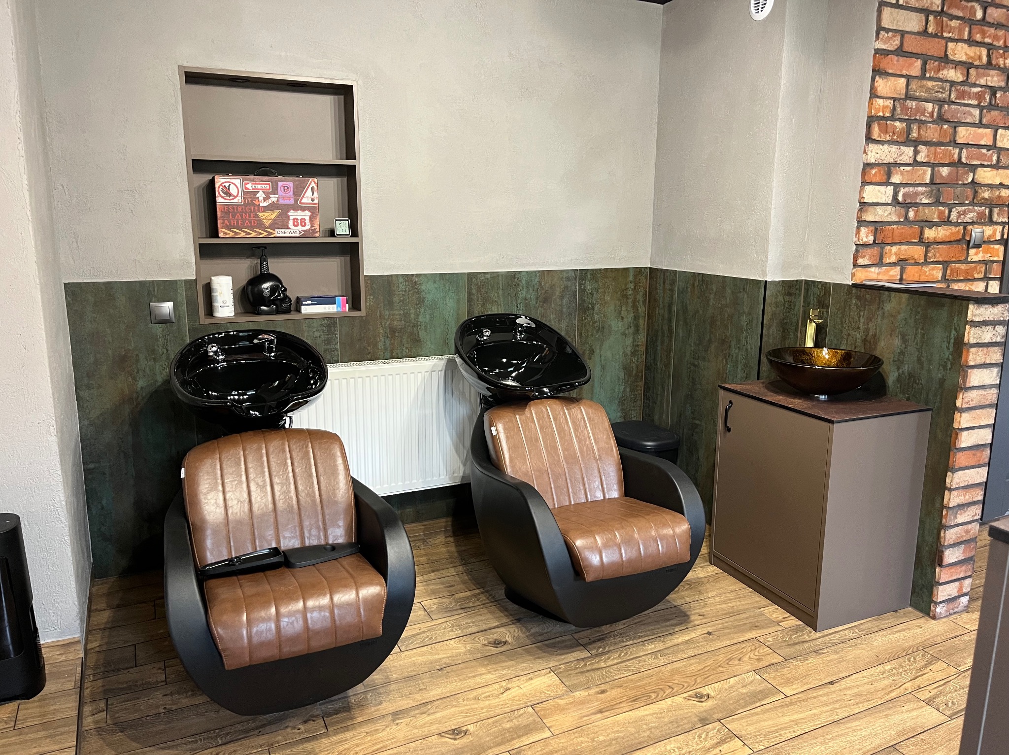 Dwa fotele fryzjerskie z umywalkami w salonie, brązowe obicia, czarne konstrukcje, drewniana podłoga, ściana z cegły i półka z dekoracjami w tle.