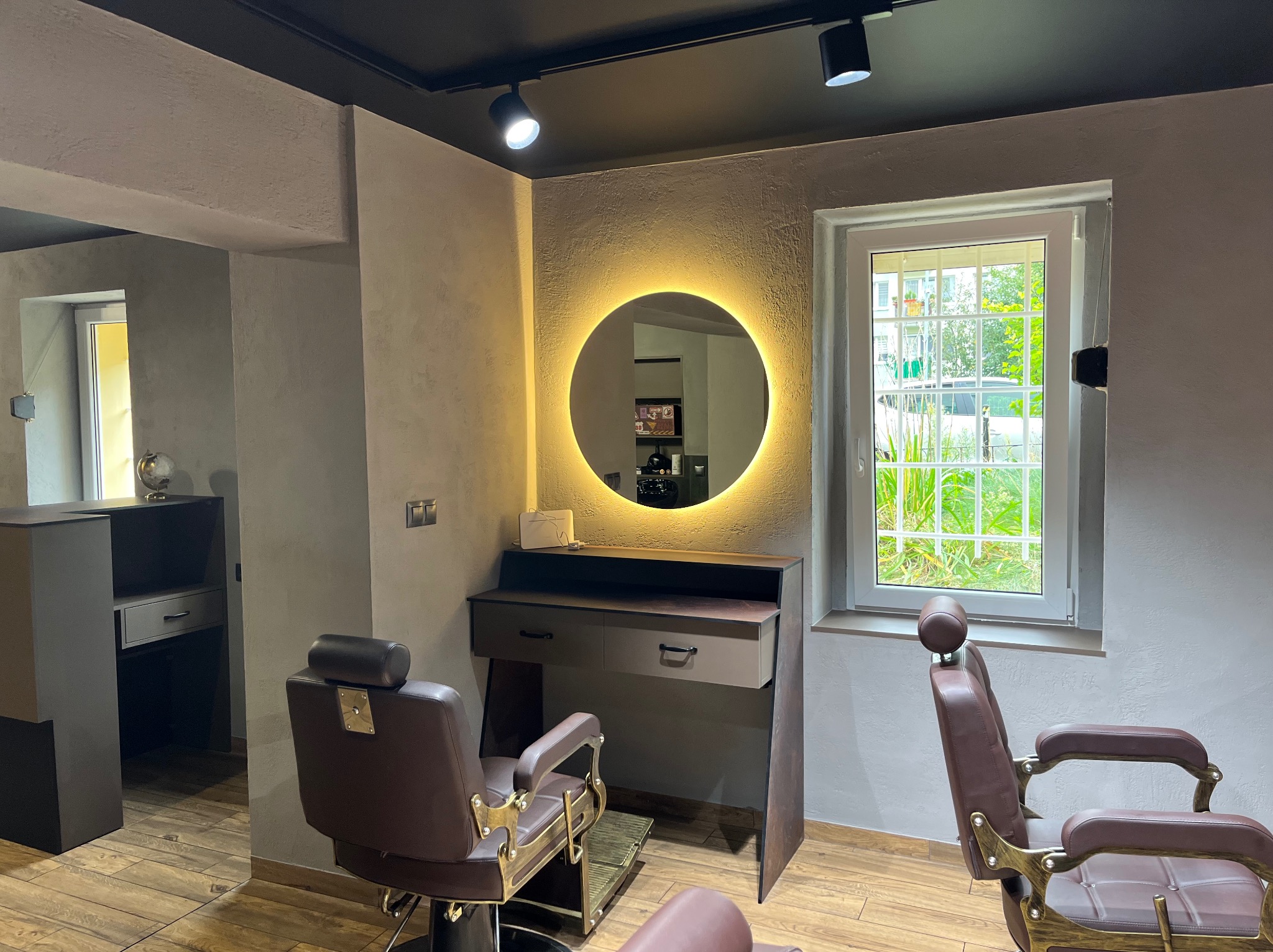 Stylowe wnętrze salonu fryzjerskiego z dwoma fotelami, toaletką z okrągłym lustrem podświetlanym LED i oknem z widokiem na zieleń. Nowoczesny design z elementami retro.