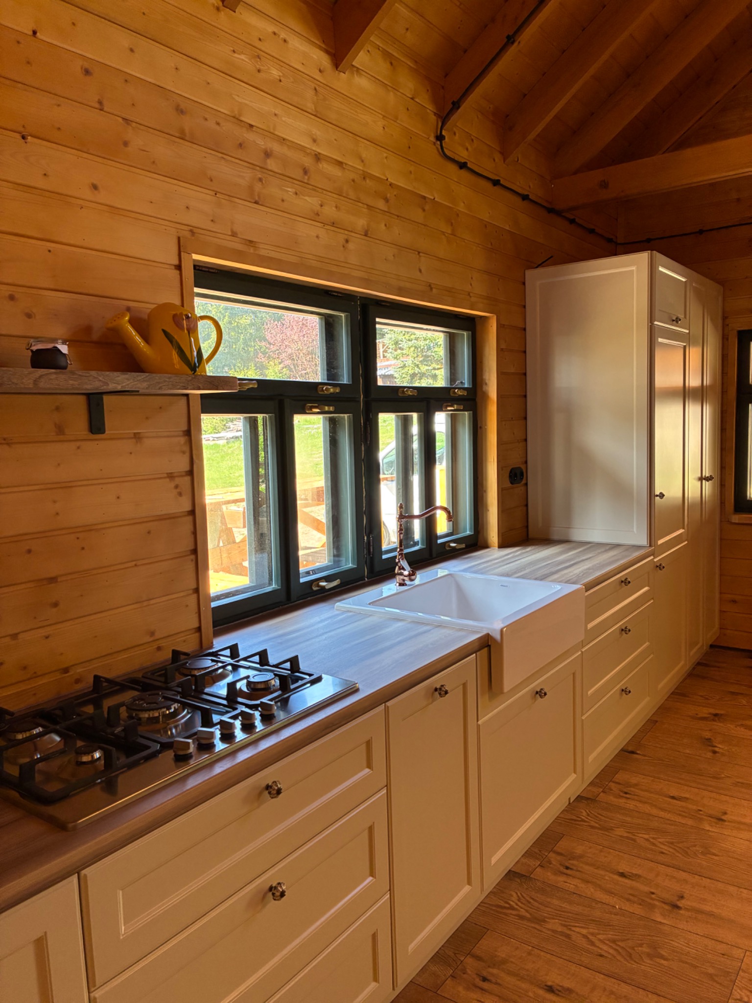 Drewniana kuchnia z oknem na ogród. Kremowe szafki z ozdobnymi uchwytami, biały zlew farmerski i płyta gazowa. Żółta konewka na półce dodaje koloru.
