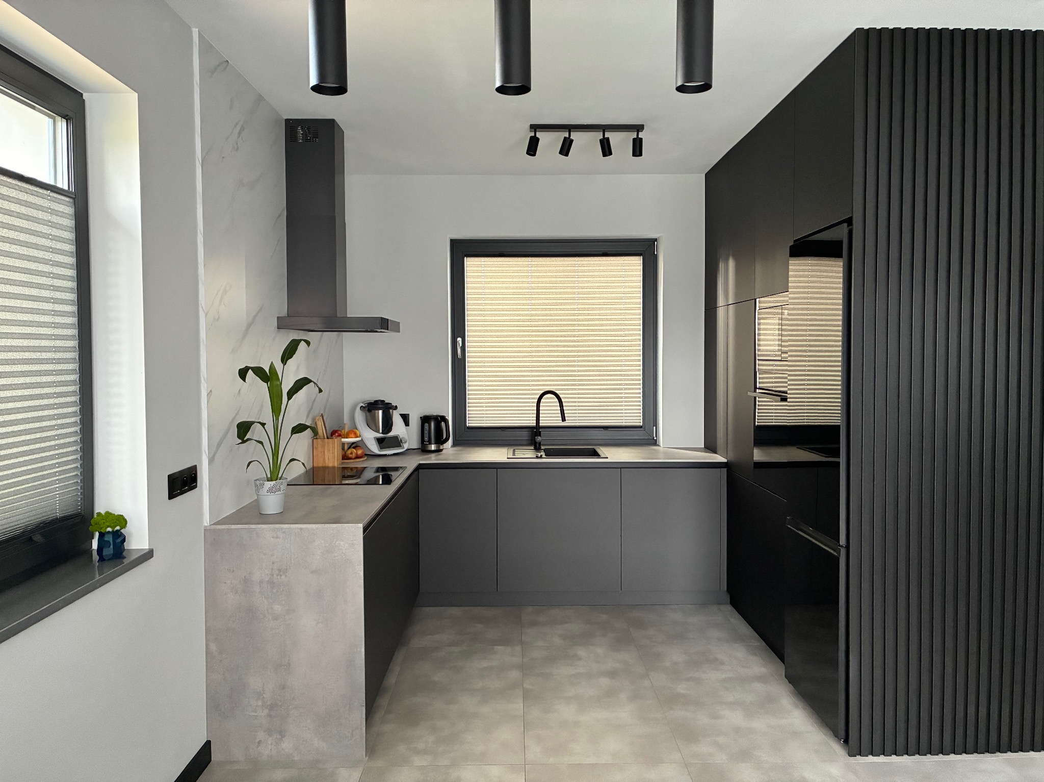 Nowoczesna kuchnia w odcieniach szarości i czerni z minimalistycznym oświetleniem punktowym i rośliną doniczkową na blacie. Fronty szafek gładkie, bez uchwytów.