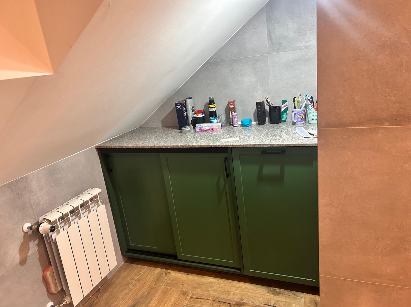 Zielona szafka pod skosem dachu z blatem w łazience, na blacie kosmetyki i akcesoria higieniczne. Grzejnik obok szafki. Ujęcie z dołu, widoczne płytki na ścianie.