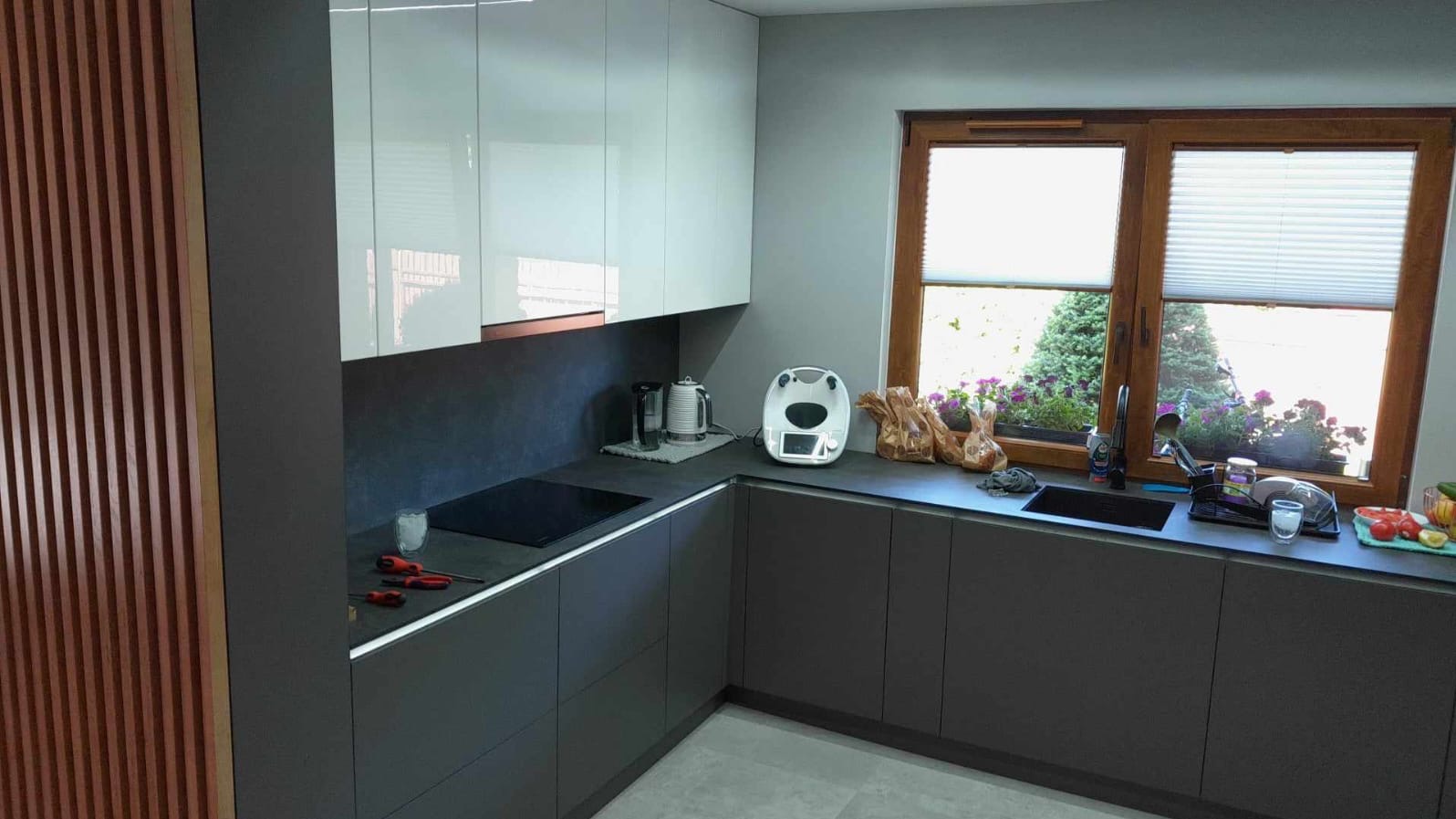 Nowoczesna kuchnia z szarymi szafkami i białymi górnymi frontami. Na blacie czajnik, płyta indukcyjna i akcesoria kuchenne. Okno z widokiem na zieleń.