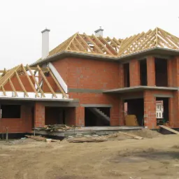 Surowy dom z czerwonej cegły z drewnianą konstrukcją dachu w trakcie budowy, widoczne kominy i schody. Plac budowy w niepogodny dzień.